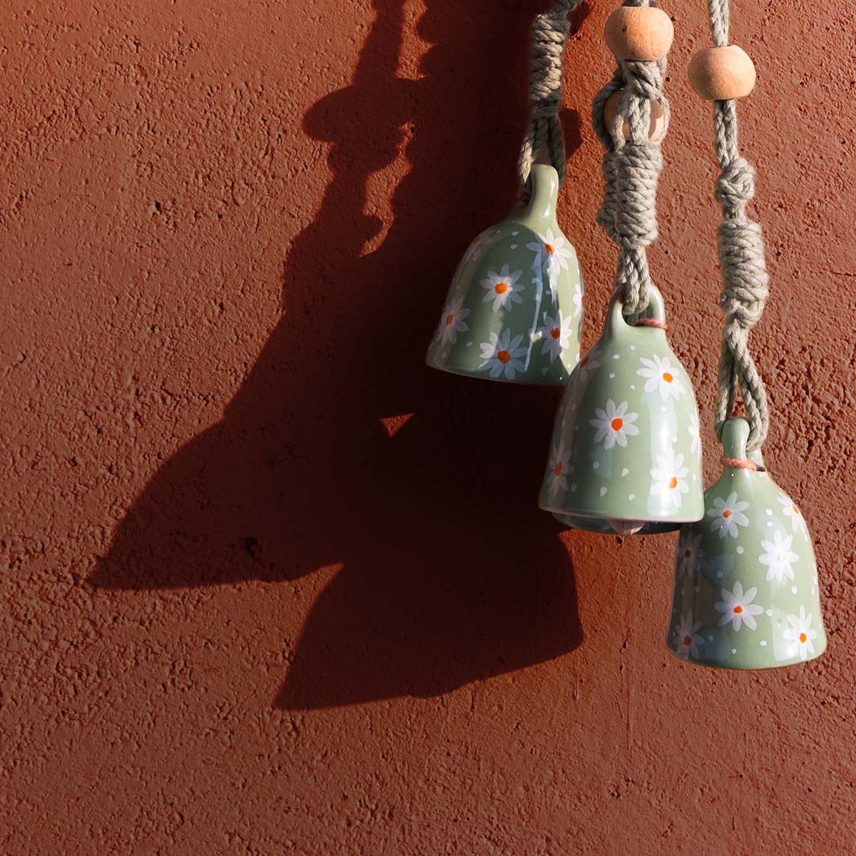 Three green ceramic macrame bells with floral patterns hanging against a red-brown wall.